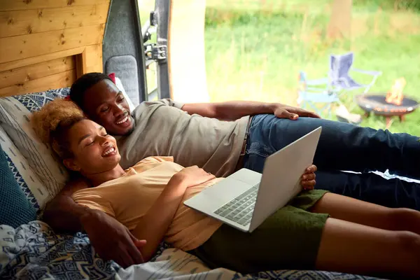 Couple Enjoying Camping In Countryside Lying Inside RV Streaming Movie On Laptop Together