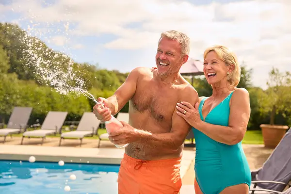 Senior Couple On Holiday In Swimming Costumes Opening Champagne By Hotel Swimming Pool