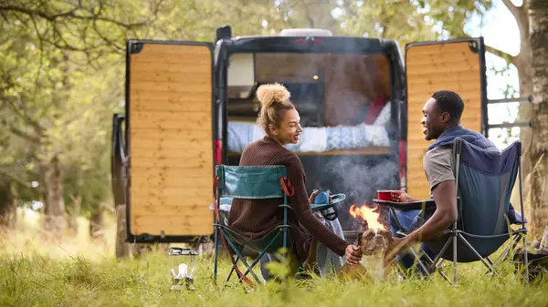 Rear View Of Couple Camping In Countryside With RV Drinking Coffee By Outdoor Fire With Pet Dog