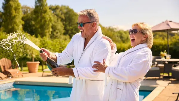 Senior Couple Holiday Wearing Robes Opening Champagne Hotel Swimming Pool — Stock Photo, Image