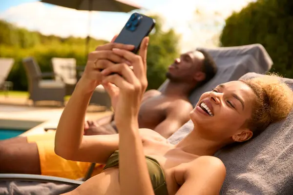 Couple On Holiday Wearing Swimming Costumes Looking At Mobile Phone On Lounger By Pool
