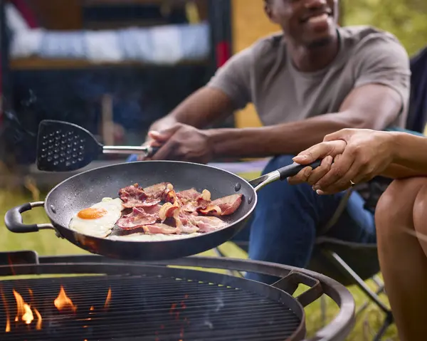 Close Up Of Couple Camping In Countryside With RV Cooking Bacon And Eggs For Breakfast On Fire