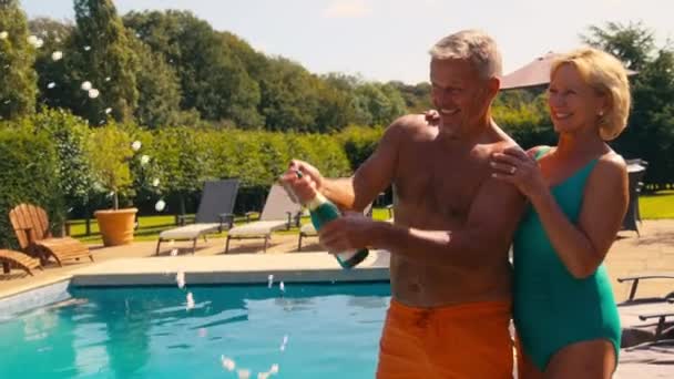 シニア引退したカップルは 夏のスパ休日にラウンジャーでホテルのプールでシャンパンを開いて吹き飛ばすことを祝うスイミングコスチュームを着て スローモーションで撮影 — ストック動画