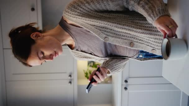 ソーシャルメディアや携帯電話のメッセージをチェックするキッチンカウンターで立っているコーヒーのカップで休憩を取る若い女性の垂直ビデオ スローモーションで撮影 — ストック動画