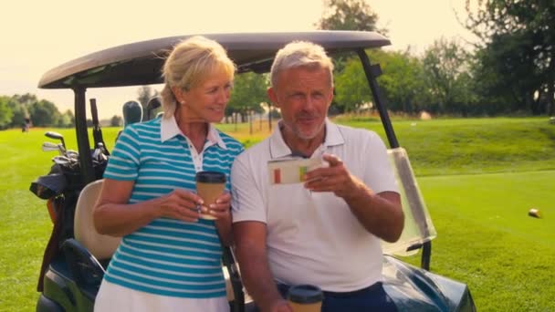 スコアカードを一緒に見てゴルフのラウンド中にテイクアウトコーヒーを持っているバギーの隣のシニア引退カップル スローモーションで撮影 — ストック動画