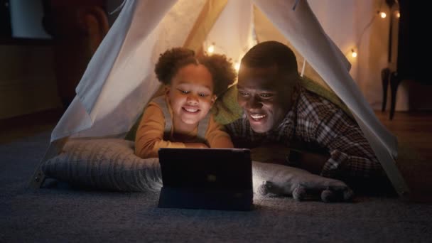 父亲和女儿躺在家里的帐篷或营地里观看或流连到数字平板电脑上 动作缓慢 — 图库视频影像