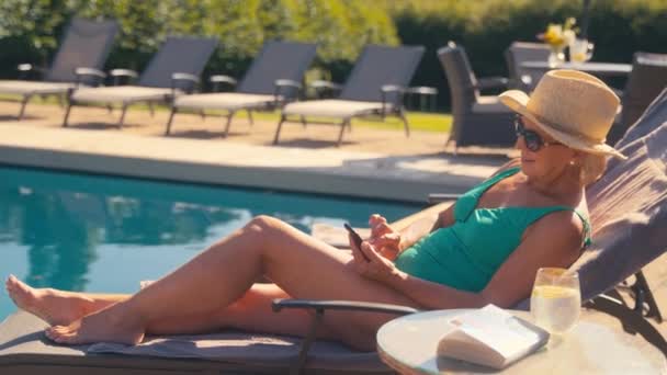 スイミングコスチューム サンハット サングラスを着用した上級退職女性は 夏の休日に携帯電話でソーシャルメディアを見るラウンジャーでホテルプールでリラックス スローモーションで撮影 — ストック動画