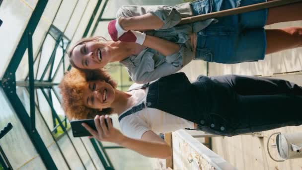 携帯電話でセルフィーのためにポーズする自宅で温室で働く2人の女性の友人の垂直ビデオ肖像画 スローモーションで撮影 — ストック動画