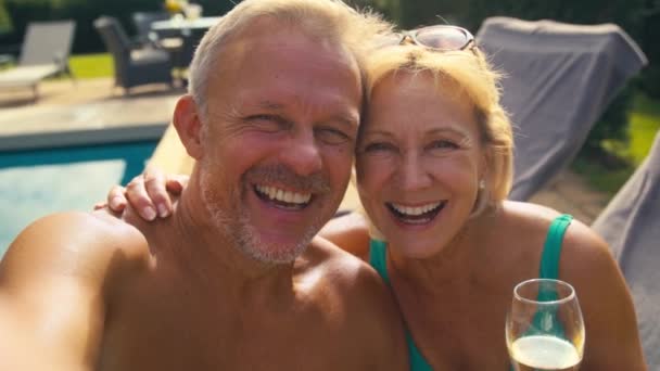 夏の休日にラウンジャーでホテルのプールでリラックスして乾杯する携帯電話でセルフィーのためにポーズを着た上級引退したカップルのPovショット スローモーションで撮影 — ストック動画