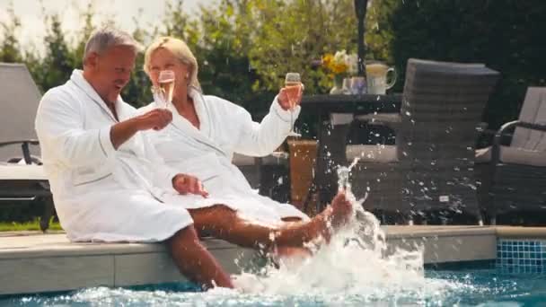 シニア引退したカップルは シャンパンを飲むローブを着て 夏のスパ休日にホテルのスイミングプールでスプラッシュ スローモーションで撮影 — ストック動画