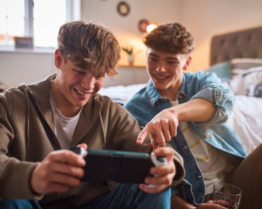 Two Teenage Boys At Home In Bedroom Together Playing On Handheld Computer Gaming Device Together clipart