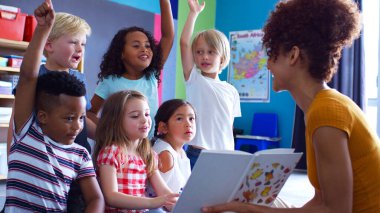 Elementary Pupils Raising Hands To Answer Question As Female Teacher Reads Story In Classroom clipart