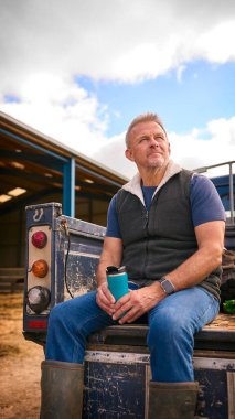 Mature Male Farm Worker Sitting On Tailgate Of Off Road Farm Vehicle Holding Hot Drink In Cup clipart