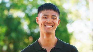 A young man stands confidently in a park, smiling broadly while enjoying the warm sunshine. The vibrant greenery of trees in the background enhances the cheerful atmosphere, creating a relaxed and inviting environment.  clipart