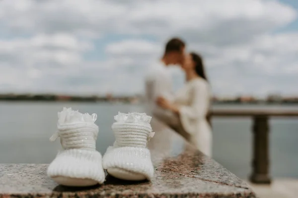 Family shoes in the sand with pregnant woman in the background kissing her partner. Selective focus on shoes in foreground