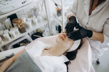 Kız yüzüne masaj yapan ve modern güzellik salonundaki kameraya bakan kadın güzellik uzmanının en iyi görüntüsü. Kapalı gözlü, tıbbi şapkalı genç Asyalı kadın müşteri. Vücut ve cilt bakımı kavramı