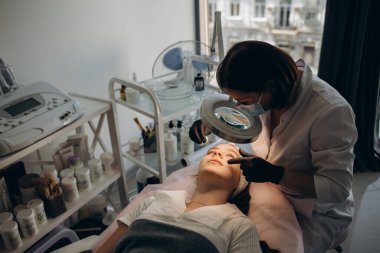 Orta yaşlı kadın kozmetikçi, özel büyüteç kullanarak yaşlı bir kadın için kaş cımbızı yapıyor.