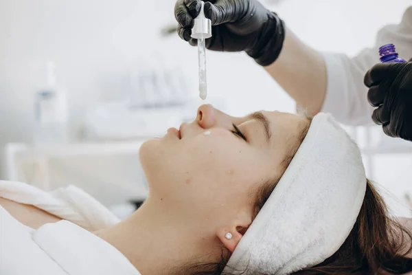 Cropped image of female beautician dripping moisturizing oil with dropper on face of girl in beauty salon. Blurred young asian woman client with closed eyes wearing medical cap. Body and skin care