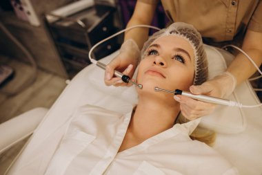 A doctor cosmetologist makes a microcurrent facial therapy to a young woman with a device in a beauty wellness salon.Cosmetology and professional skin care.