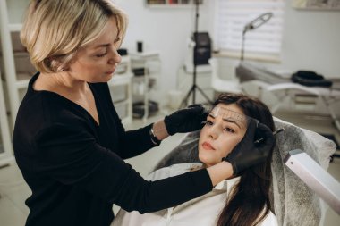 Permanent make-up for eyebrows of beautiful woman with thick brows in beauty salon. Closeup beautician doing tattooing eyebrow. Professional makeup and cosmetology skin care.