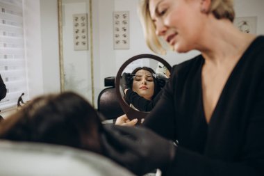 Professional beautician master of permanent makeup in protective medical mask at cosmetology cabinet makes tattooing eyes beautiful girl, concept of beauty salon and permanent make-up