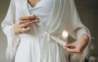 Close-up photo of a bride in silk dressing gown holding wedding dress infront of the window. Wedding morning, bride's preparations.