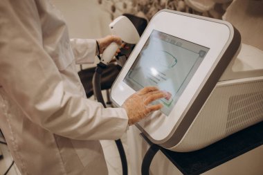 Aesthetic body treatment. Close up back side portrait of cosmetologist adjusting settings on monitor for laser hair removal device while young woman lying on couch.