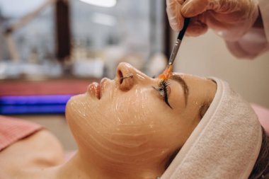 Beautician working with real woman client. Woman getting facial peeling by cosmetologist. Beautician applying clay face mask on woman face. Cosmetologist applying mask on clients face in spa salon.