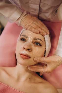 Beautician working with real woman client. Woman getting facial peeling by cosmetologist. Beautician applying clay face mask on woman face. Cosmetologist applying mask on clients face in spa salon.