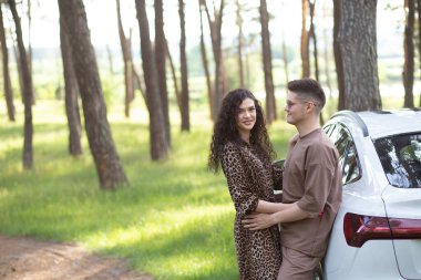 Genç modern çift doğada piknik yapıyor, park halindeki beyaz elektrikli arabanın yanında oturuyorlar.