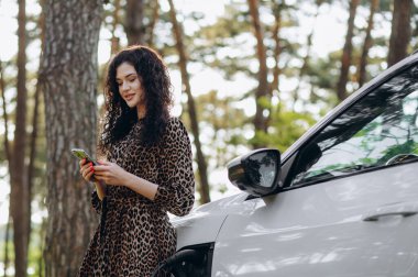 Beklerken akıllı telefon kullanmak. Elektrikli arabalardaki kadın gündüz şarj istasyonunda. Yepyeni bir araç..