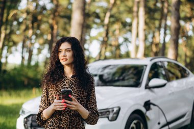 Güneşli alışveriş merkezinin otoparkında elektrikli araba beklerken telefon ekranına bakan mutlu beyaz kadın. Yaşam tarzı ve ekoloji kavramı