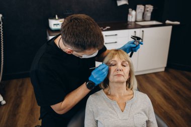Charming older woman getting face lift shot over light studio background. Unrecognizable beautician or plastic surgeon making beauty injection to attractive senior female patient