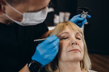 Charming older woman getting face lift shot over light studio background. Unrecognizable beautician or plastic surgeon making beauty injection to attractive senior female patient