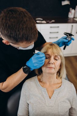 Plastic surgeon in protective gloves injecting face filler to senior woman over light studio background. Lovely mature lady getting anti-aging procedure. Aesthetic medicine concept