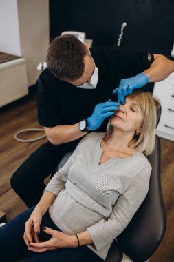 Plastic surgeon in protective gloves injecting face filler to senior woman over light studio background. Lovely mature lady getting anti-aging procedure. Aesthetic medicine concept