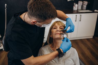 Plastic surgeon in protective gloves injecting face filler to senior woman over light studio background. Lovely mature lady getting anti-aging procedure. Aesthetic medicine concept