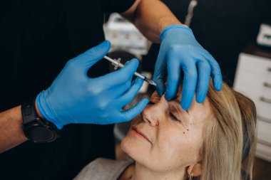 Plastic surgeon in protective gloves injecting face filler to senior woman over light studio background. Lovely mature lady getting anti-aging procedure. Aesthetic medicine concept