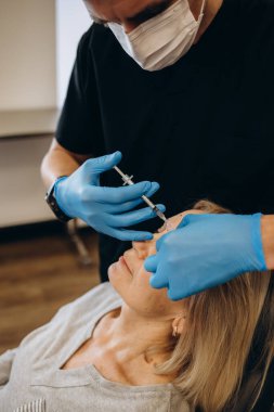 Plastic surgeon in protective gloves injecting face filler to senior woman over light studio background. Lovely mature lady getting anti-aging procedure. Aesthetic medicine concept