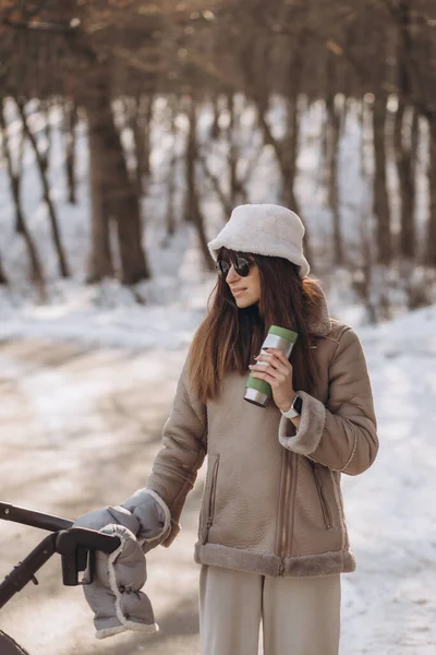 Stylish young mom with a cup of coffee on a walk with a baby carriage in winter