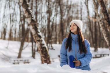 Happy young woman drinks hot tea from thermos cup during walk in winter forest.