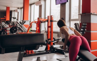 Güçlü bir vücut geliştirici spor salonundaki bir bankta eğilir ve dambıllarla antreman yapar..