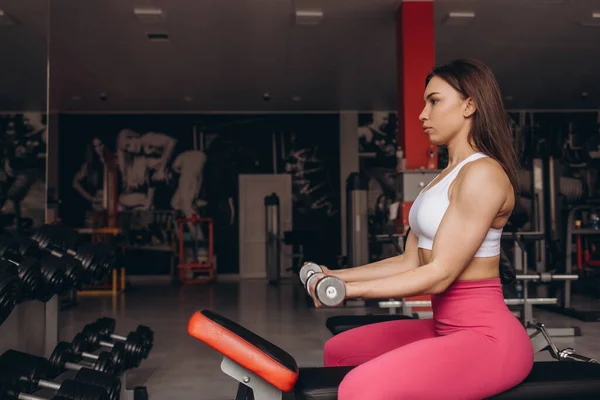 stock image Smiling sport woman training biceps with dumbbell in hand while sitting on bench in gym