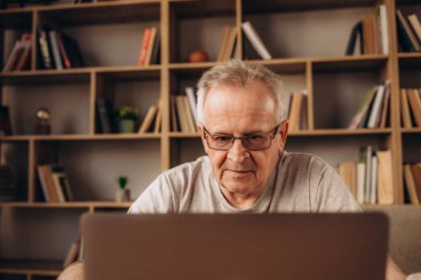 Laptop ile evde çalışan üst düzey adam. Evde bilgisayarı kullanan ihtiyar sandalyede oturan ve ekrana bakarak. Gözlük takan ve oturma odasında dizüstü bilgisayar üzerinde çalışan yaşlı dede.
