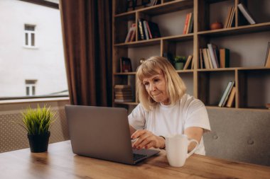 Orta yaş kıdemli kadın evde masada oturan mutlu bir yüz ayakta ve kendine güvenen bir gülümseme ile gülümseyen diş gösteren bilgisayar laptop kullanarak çalışma