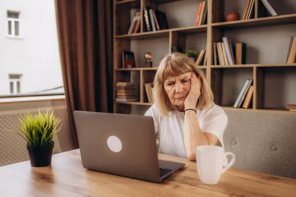 Tired senior woman sitting at table and doing self head massage. Exhausted mature lady who has headache after working on computer for a long time sitting with her eyes closed and rubbing her temples