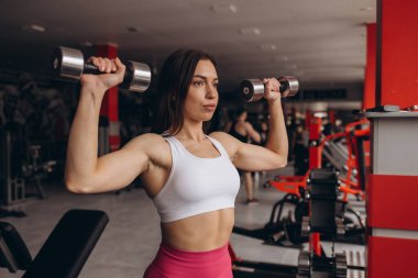 Kolları için egzersiz yapan atletik bir kadın. Spor modeli dambıllarla çalışıyor. Dambıllarla egzersiz yapan bir kadın. Fitness, egzersiz, sağlıklı yaşam ve diyet konsepti.