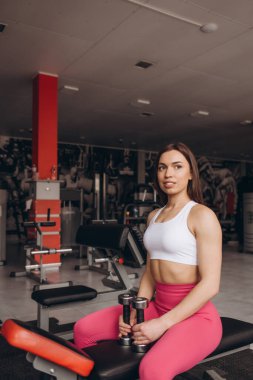 Spor yaptıktan sonra oturup dinlenen ya da spor salonunda egzersiz yapan sağlıklı genç bir kadın. Kadın spor salonunda mola veriyor ve spor giyim kuşamında suyla rahatlıyor. Fitness konsepti, Sağlıklı, Spor, Yaşam Tarzı