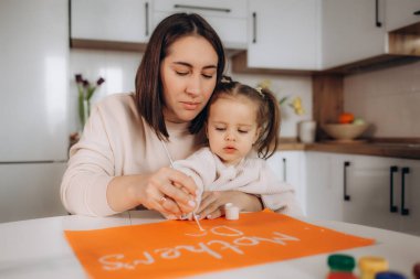 Gülümseyen küçük kız anneler günü kartı çiziyor, kırmızı bir kalp çiziyor ve annesine sarılıyor.