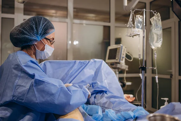 An experienced angiosurgeon operates on the leg of a patient with varicose veins. Doctor in a protective mask and an operating gown concentrates on the operation on the patient\'s leg